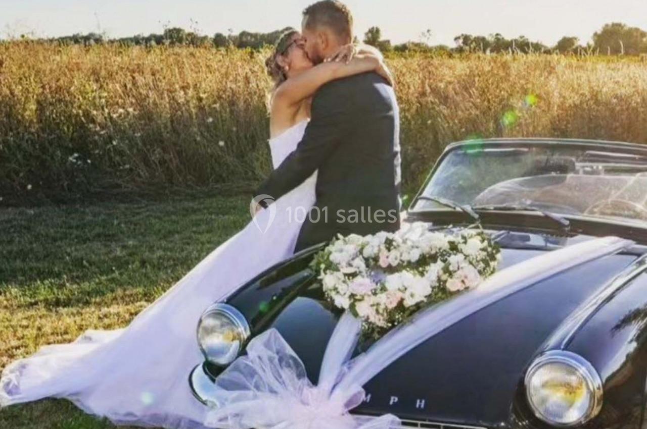 Un couple en tenue de mariage s'embrasse devant une voiture classique décorée de fleurs, dans un champ ensoleillé.
