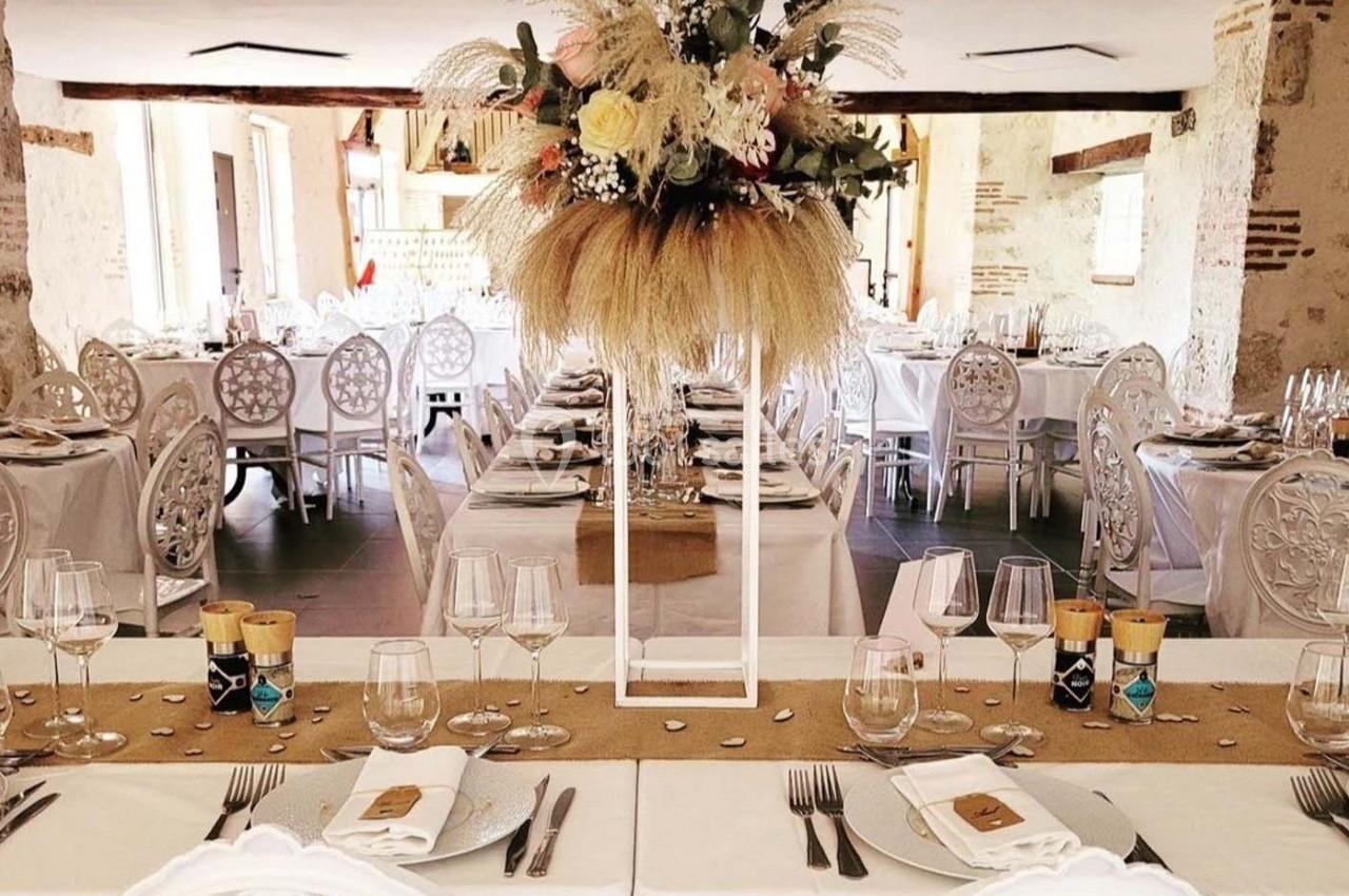 Salle de réception décorée pour un événement avec tables dressées, chaises blanches et centre de table floral élégant.