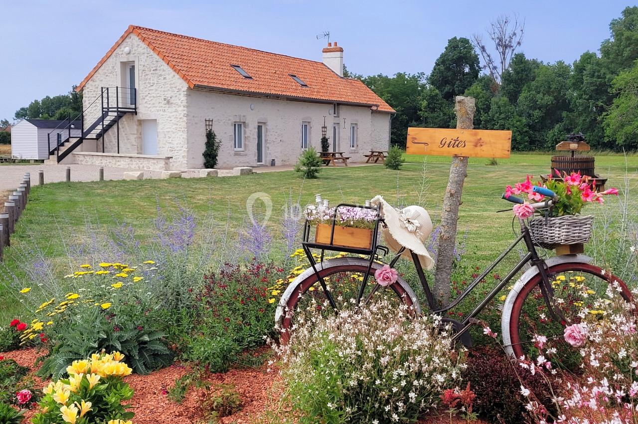 Jardin fleuri avec un vélo décoratif devant une maison en pierre à toit rouge, entourée de verdure.