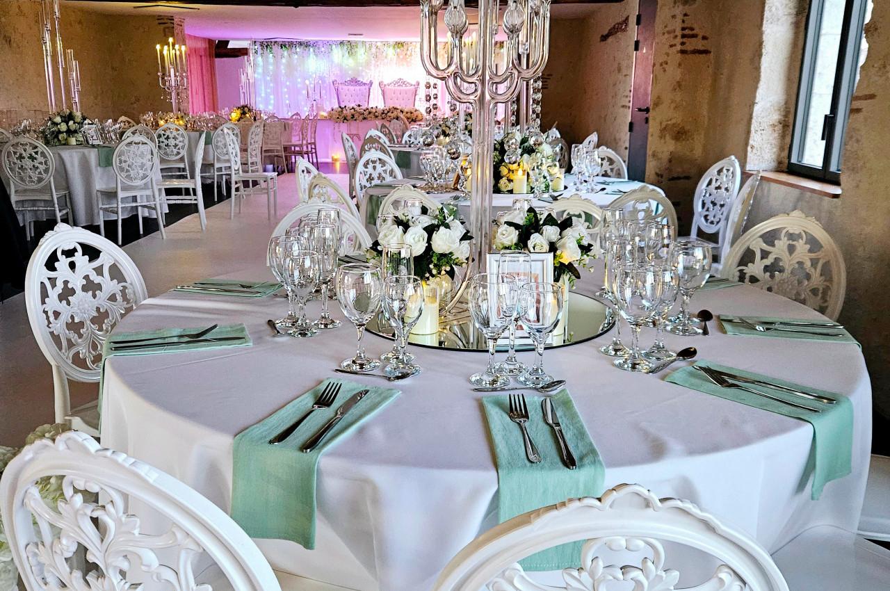 Salle de réception décorée avec des tables rondes, nappes blanches, chaises sculptées et chandeliers élégants.
