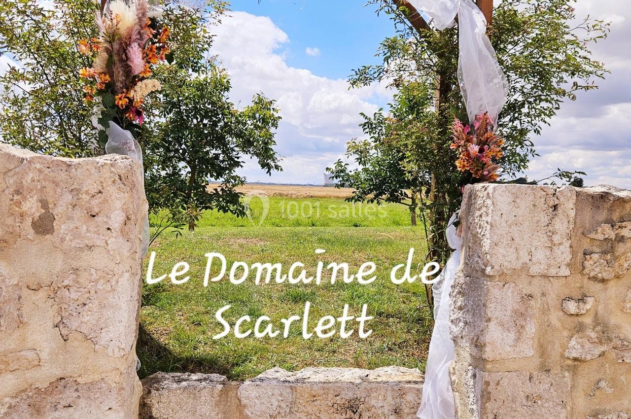 Arche en bois décorée de fleurs et de voilages blancs, encadrée par des murs en pierre, avec un champ en arrière-plan.