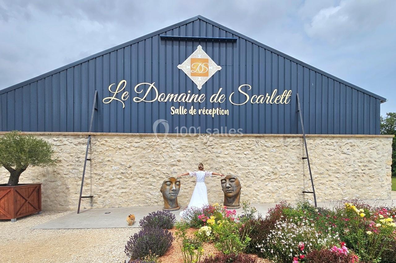 Façade d'une salle de réception en pierre et métal bleu, avec une femme en robe blanche devant des sculptures et des fleurs.