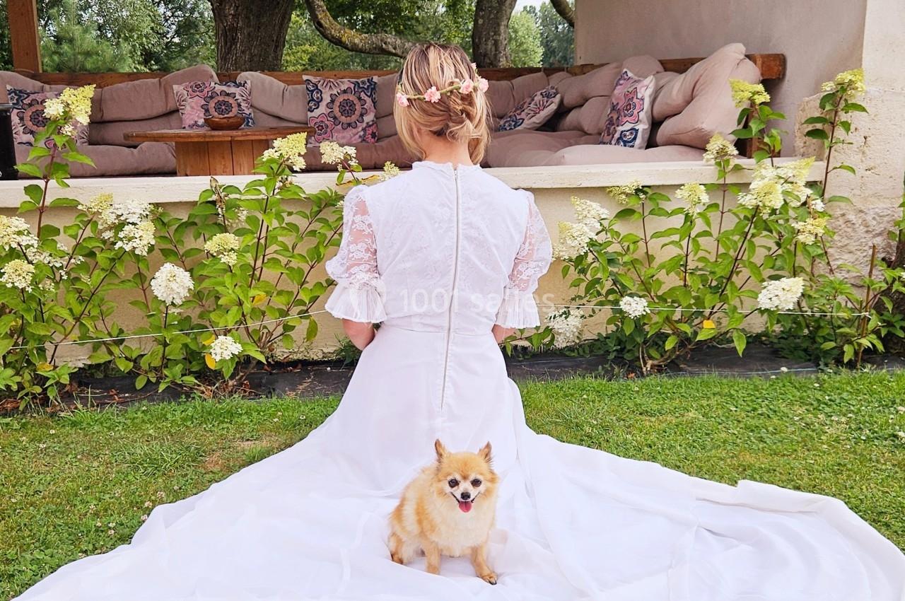 Une femme en robe blanche assise sur l'herbe, de dos, avec un petit chien devant elle.