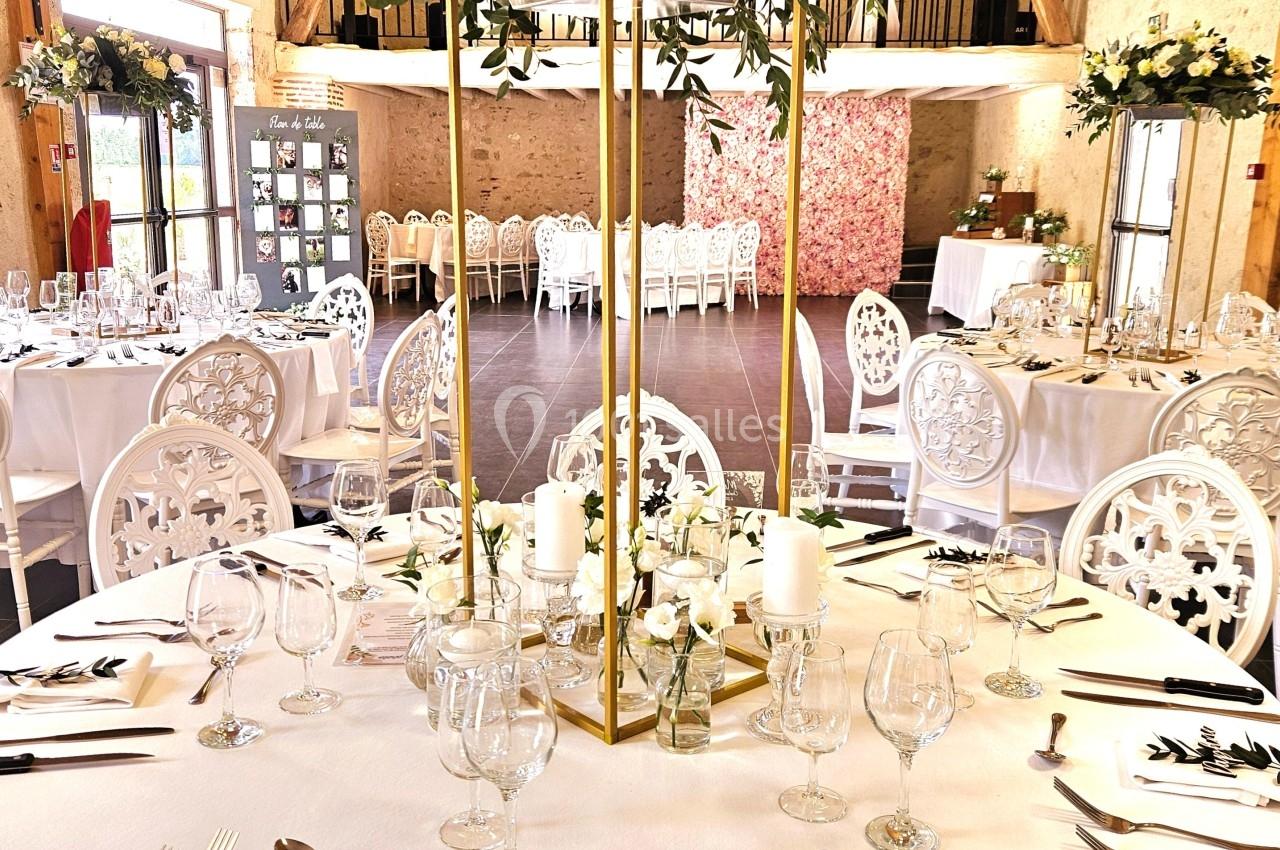 Salle de réception décorée pour un mariage avec tables rondes dressées, centres de table fleuris et chaises blanches.