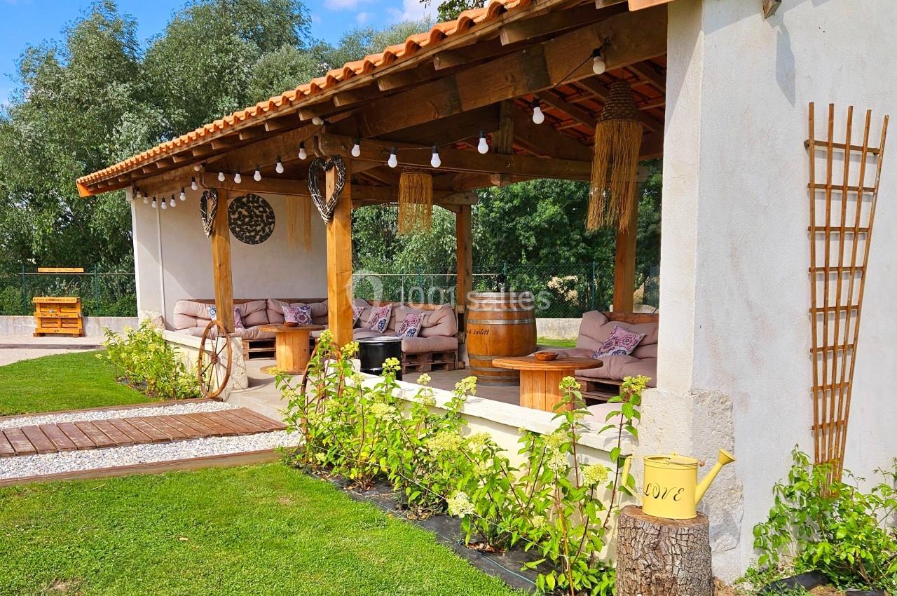 Terrasse couverte avec canapés, décor rustique et végétation, dans un jardin sous un ciel bleu.