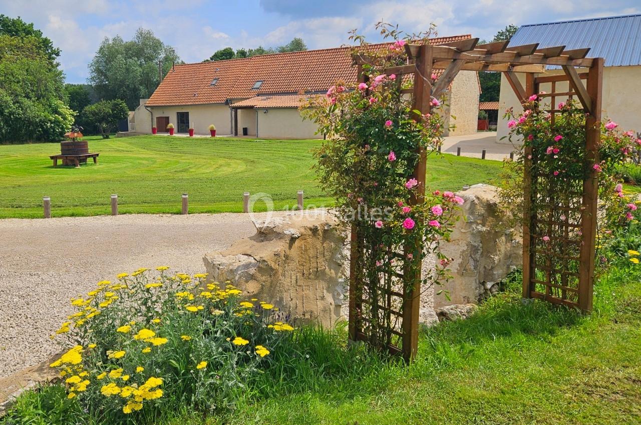 Arche en bois ornée de fleurs devant un jardin verdoyant avec une maison en arrière-plan sous un ciel dégagé.