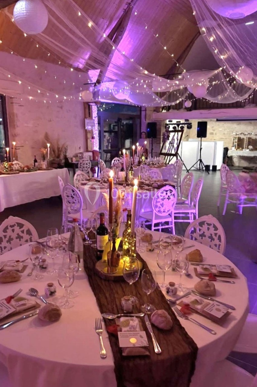 Salle de réception décorée pour un dîner, avec tables rondes dressées, bougies, guirlandes lumineuses et ambiance…