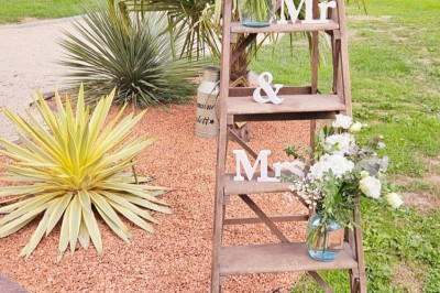 Bouquet de fleurs blanches et vertes au premier plan, avec des chaises disposées en extérieur sous un grand arbre.