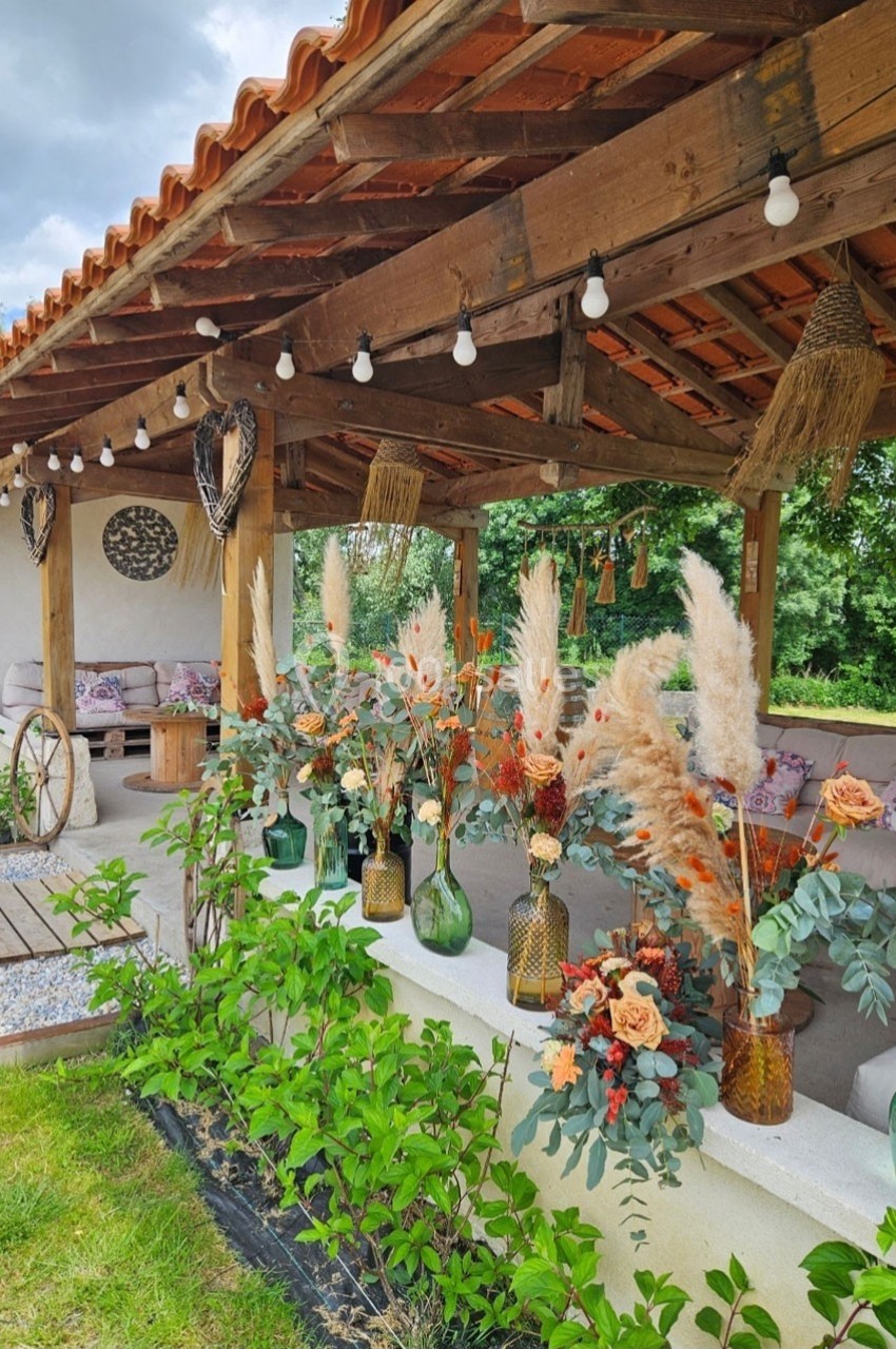 Terrasse couverte en bois décorée de vases fleuris, pampas et guirlandes lumineuses, entourée de verdure.
