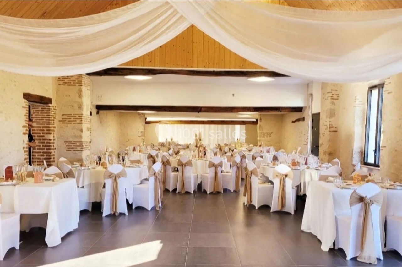 Salle de réception décorée avec des tables rondes, nappes blanches, chaises ornées de nœuds dorés et voilages au plafond.
