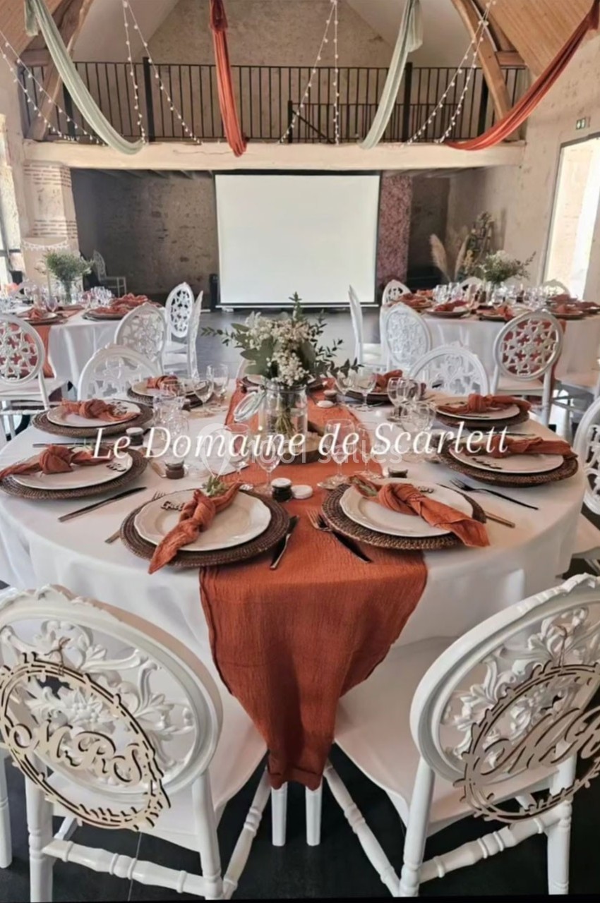Salle de réception décorée avec des tables rondes, nappes blanches, chemins de table orange et chaises blanches sculptées.