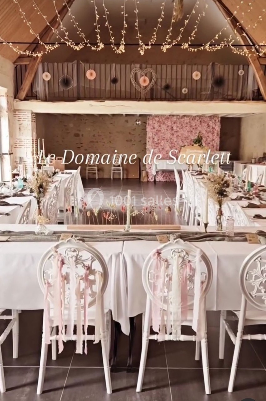 Salle de réception décorée avec des tables dressées, chaises ornées de rubans roses et guirlandes lumineuses suspendues.