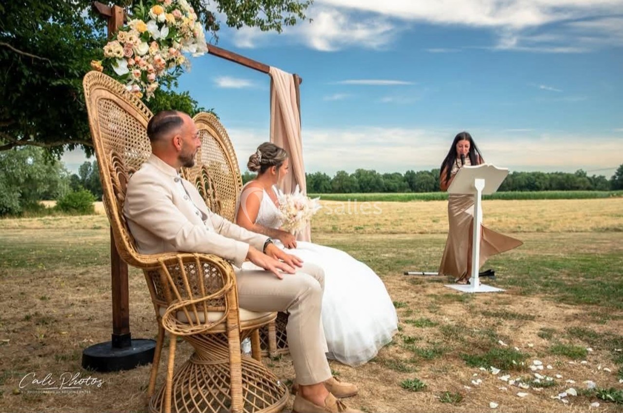 Un couple assis sur des fauteuils en osier écoute une femme prononçant un discours lors d'une cérémonie en plein air.