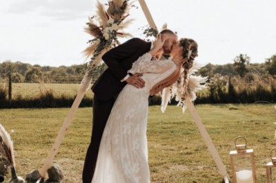 Un couple en tenue de mariage s'embrasse devant une arche décorée, dans un cadre champêtre en plein air.