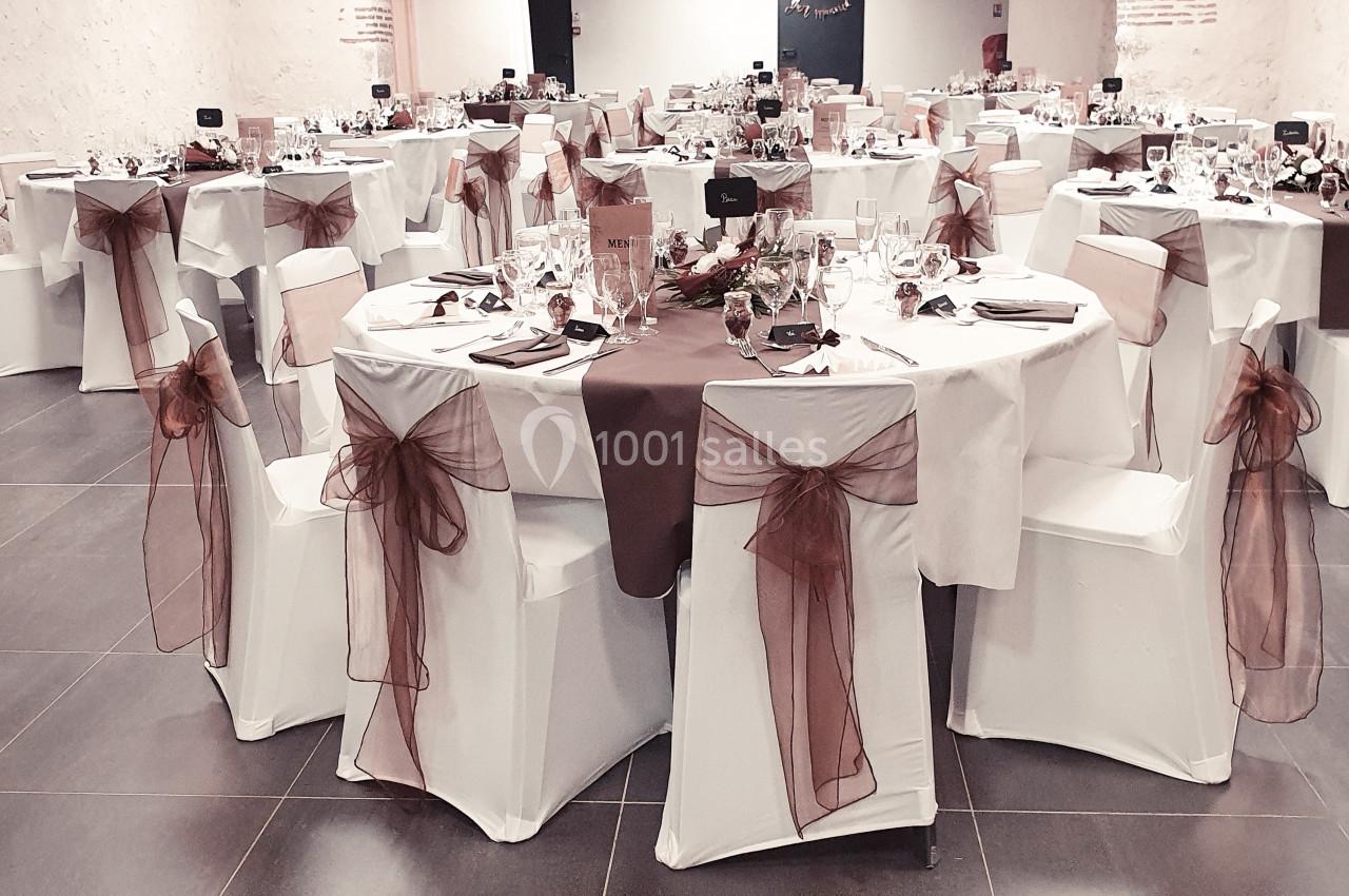 Salle de réception décorée avec des tables rondes, nappes blanches et nœuds en tissu marron sur les chaises.
