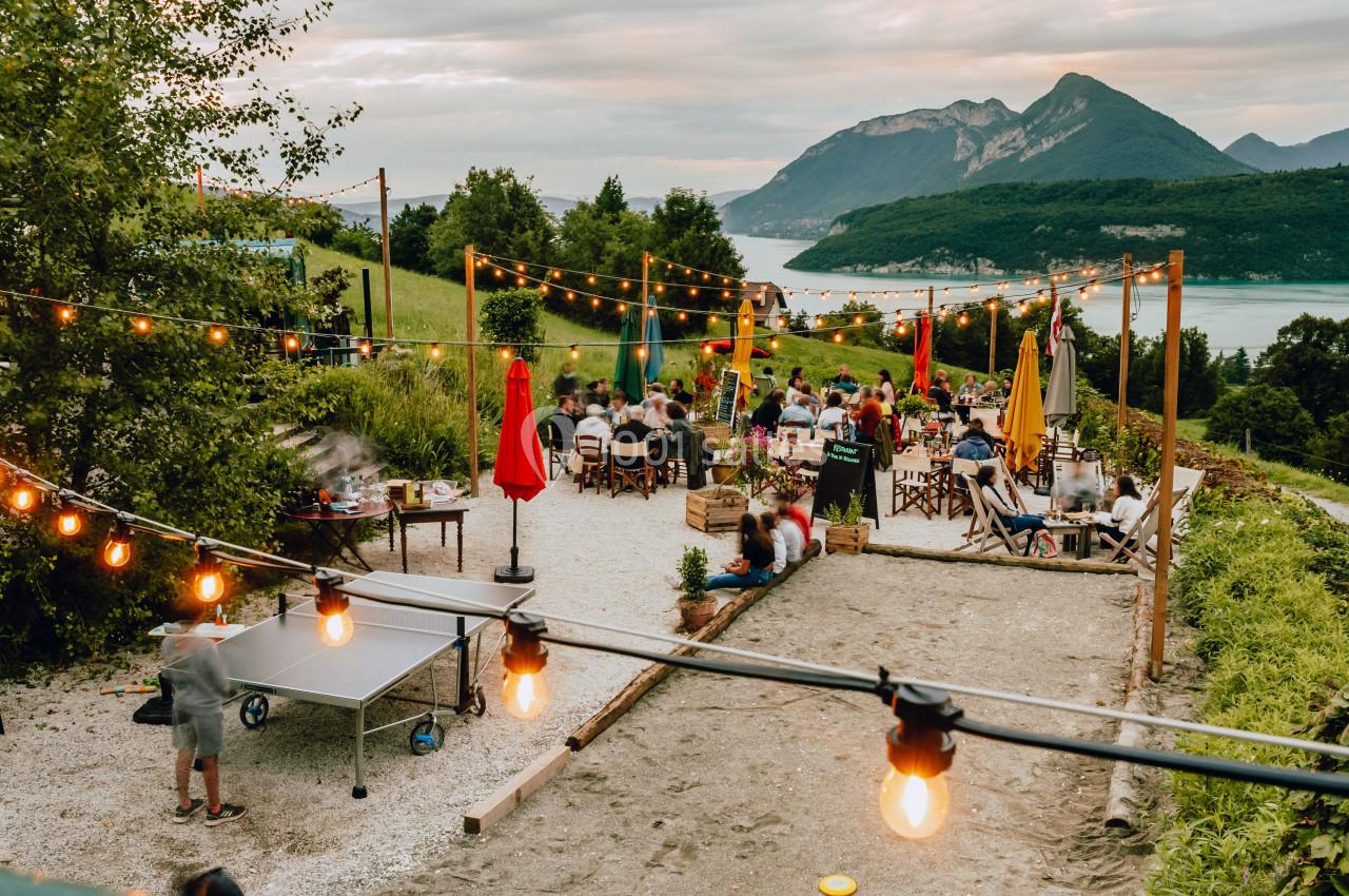 Terrasse extérieure animée avec tables, guirlandes lumineuses, vue sur un lac et montagnes en arrière-plan.