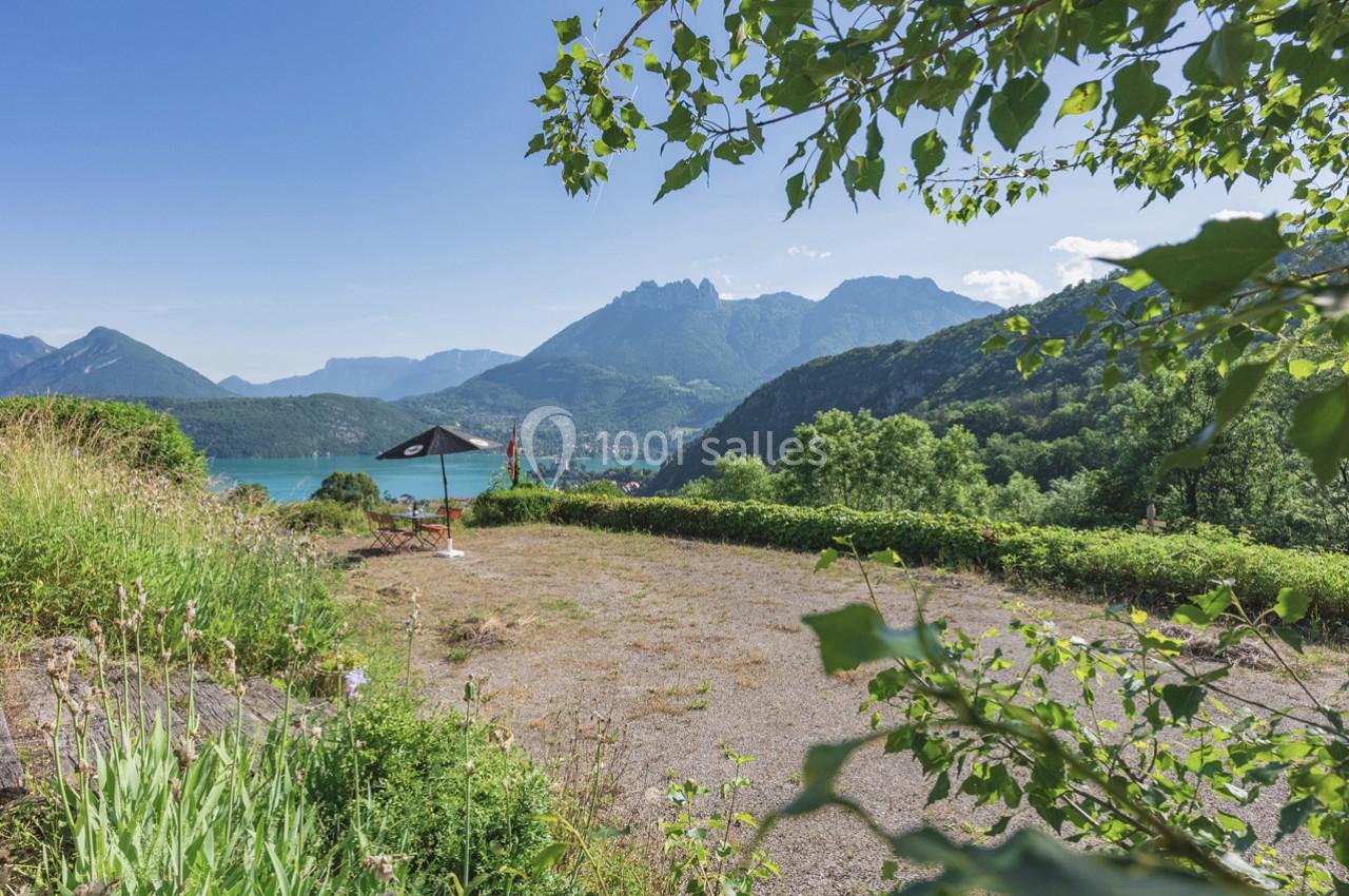 Vue sur un lac entouré de montagnes, avec un parasol noir sur une terrasse ensoleillée bordée de végétation.