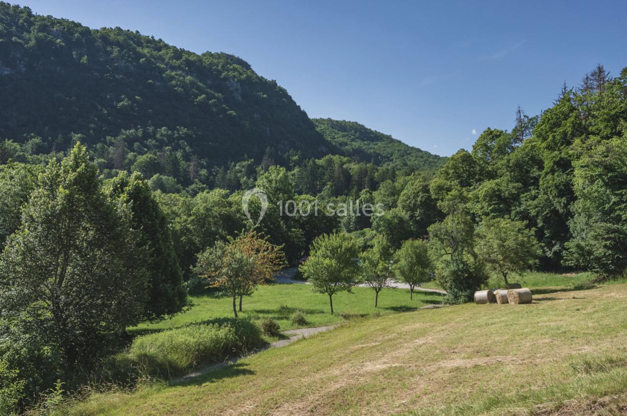 Paysage rural avec une prairie, des arbres, des bottes de foin et des collines boisées sous un ciel bleu clair.