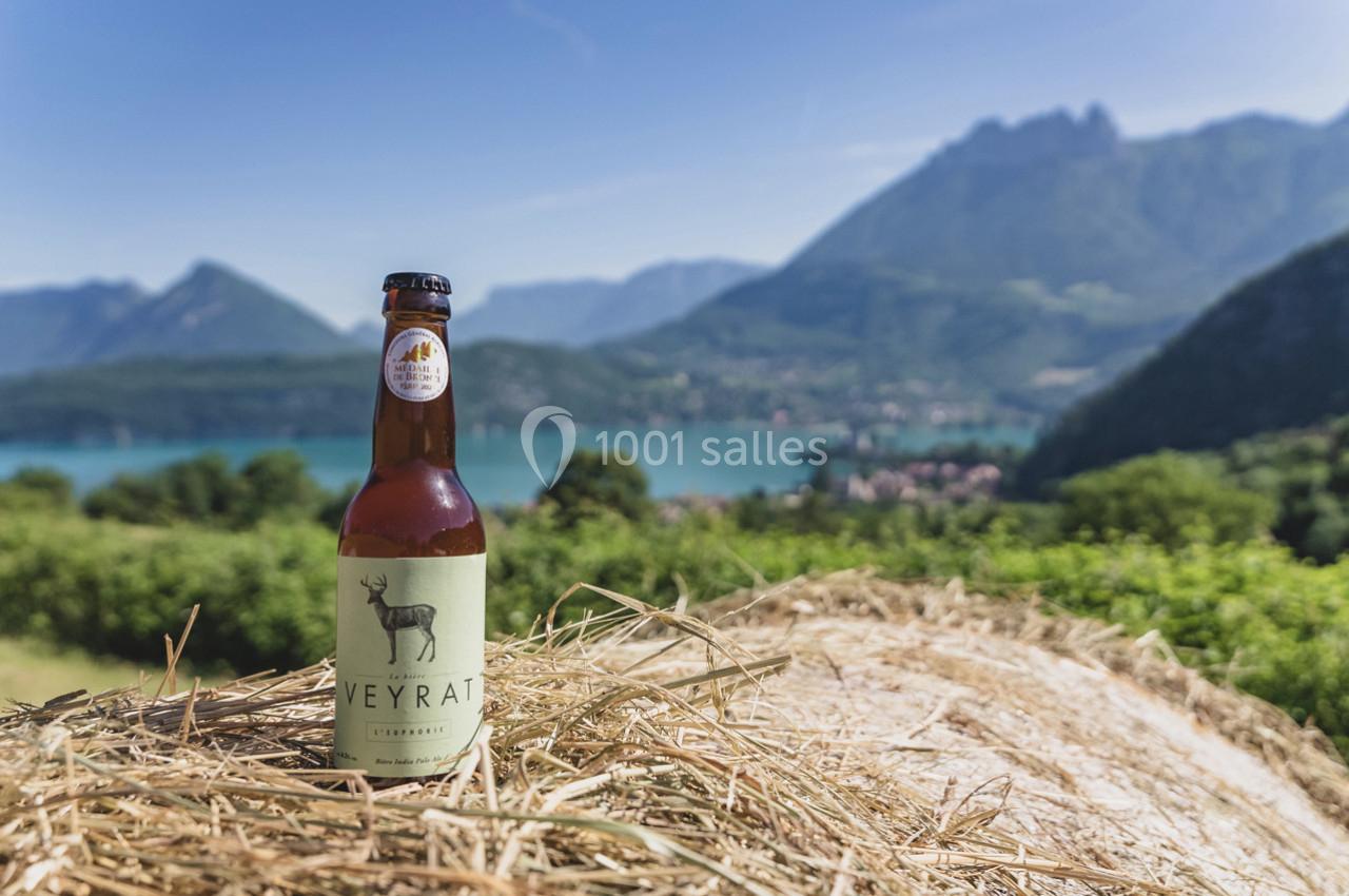 Bouteille de bière posée sur un tas de foin avec un lac et des montagnes en arrière-plan sous un ciel dégagé.