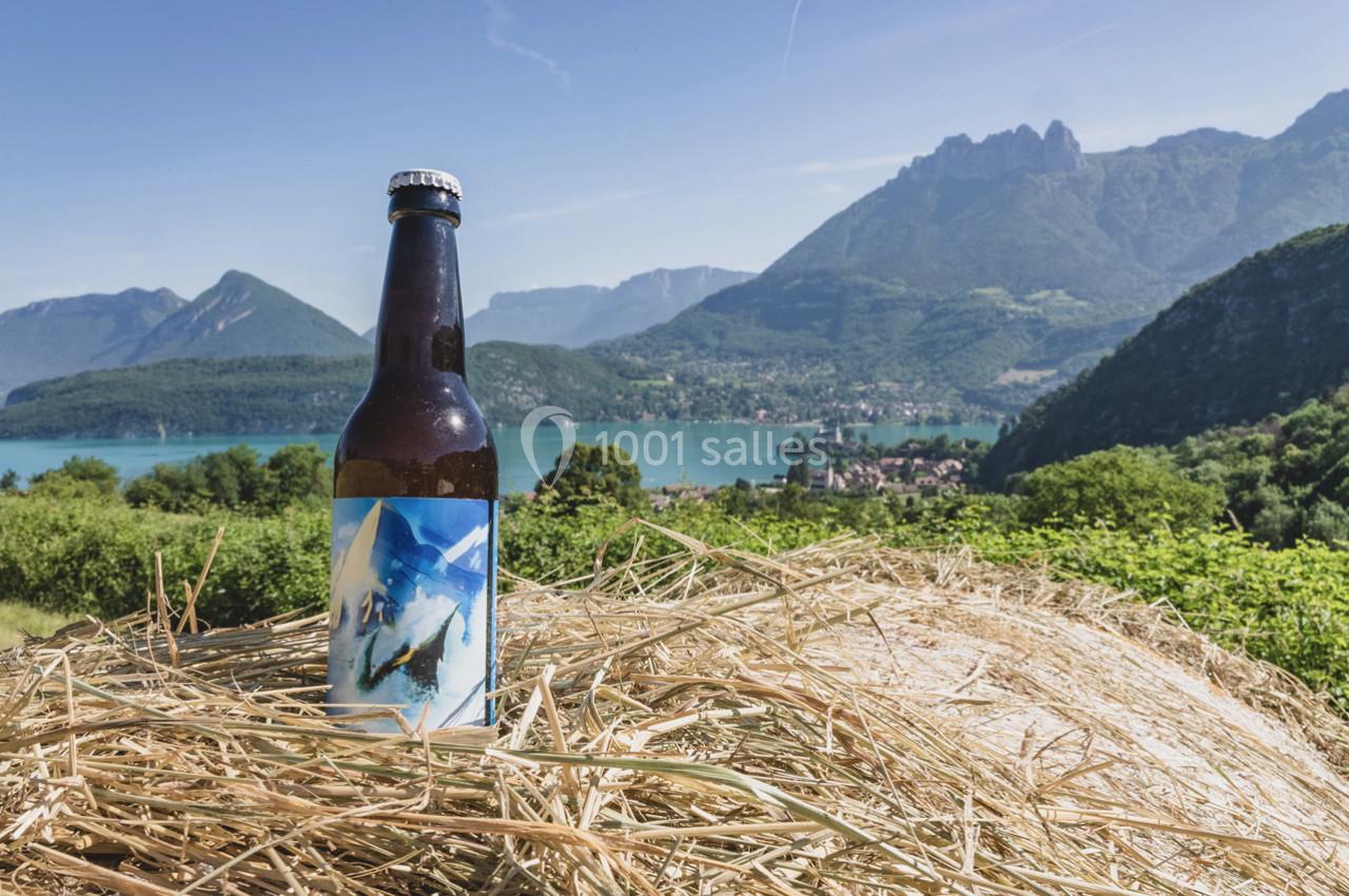 Bouteille de bière posée sur une botte de foin avec vue sur un lac entouré de montagnes verdoyantes.