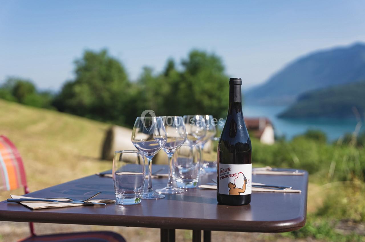 Table dressée en extérieur avec une bouteille de vin, verres et couverts, vue sur un paysage de lac et montagnes.