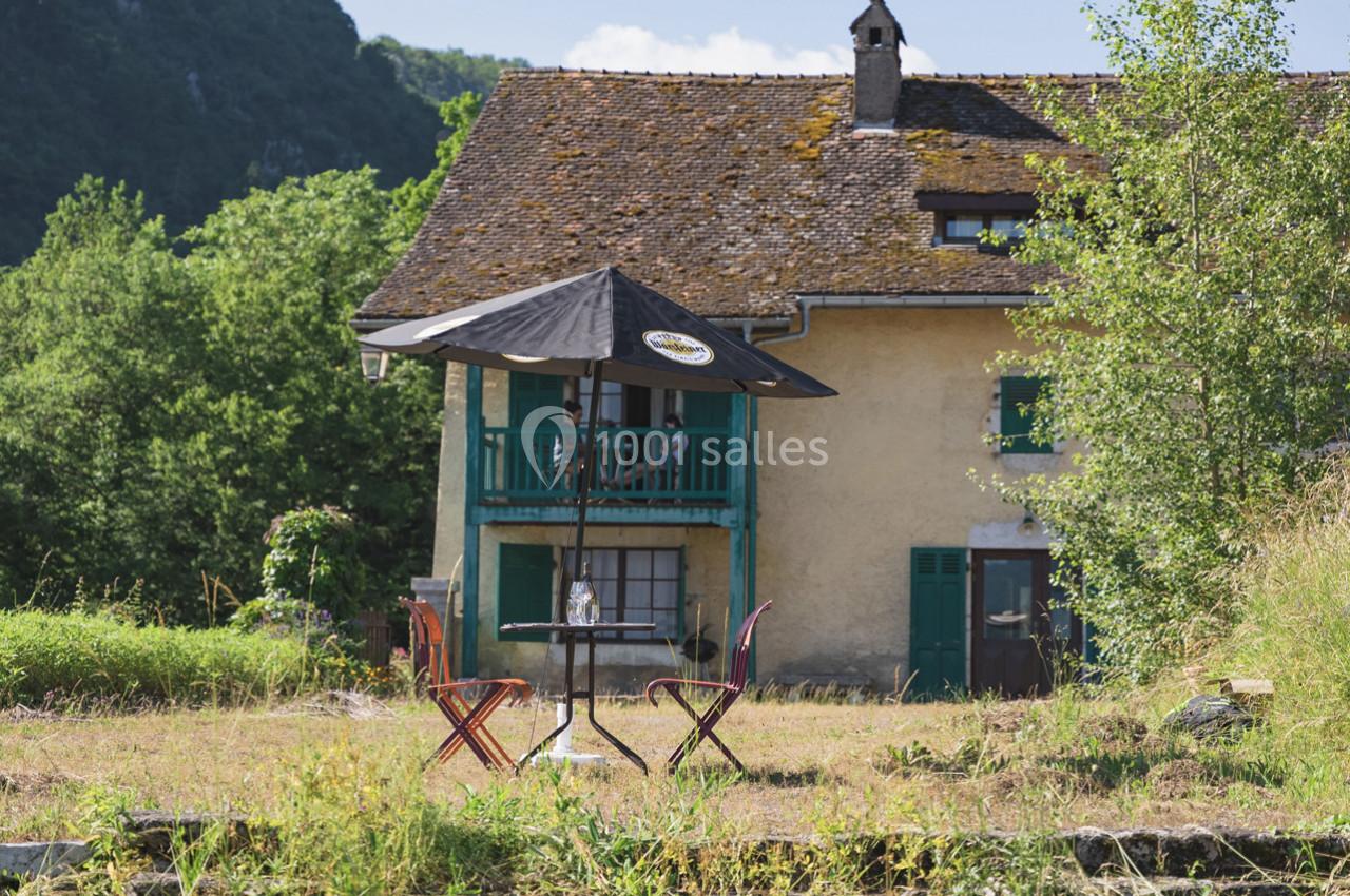 Maison ancienne avec toit en tuiles, terrasse ombragée par un parasol, entourée de verdure et collines boisées.