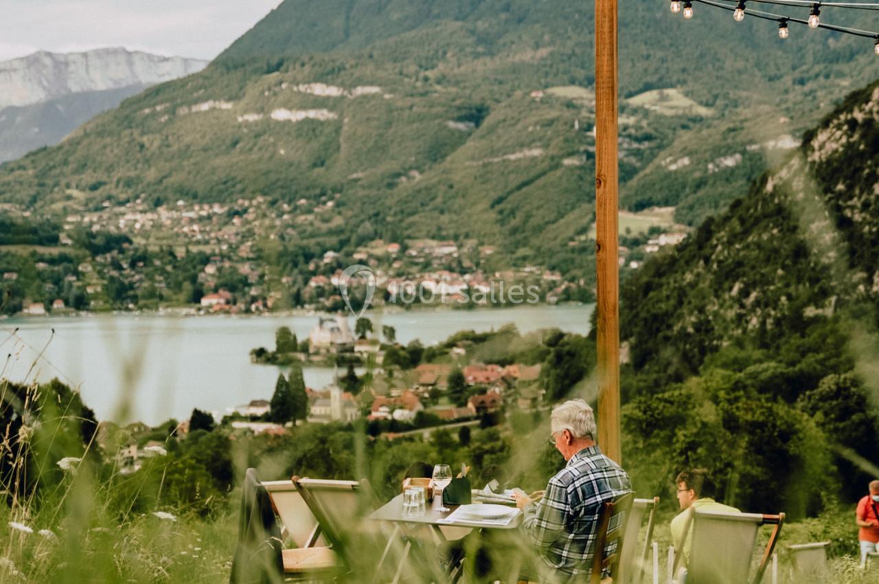 Un homme assis à une table en plein air, face à un paysage de montagne, d'un lac et d'une vallée verdoyante.