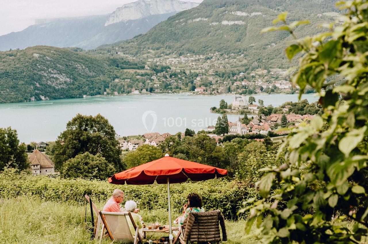 Deux personnes assises sous un parasol rouge, profitant d'une vue sur un lac entouré de montagnes et de verdure.