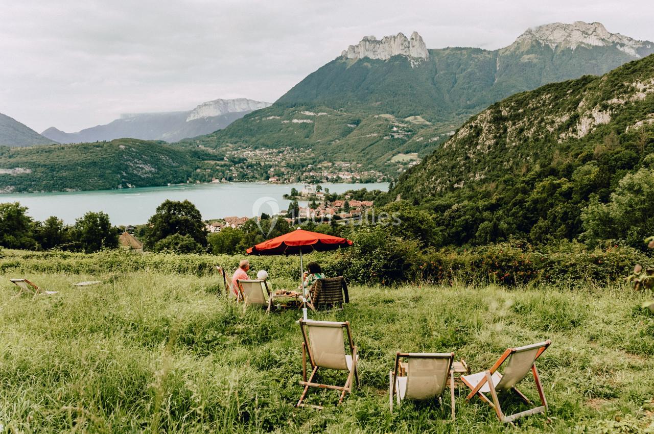 Chaises longues et parasol rouge sur une pelouse avec vue sur un lac entouré de montagnes verdoyantes.
