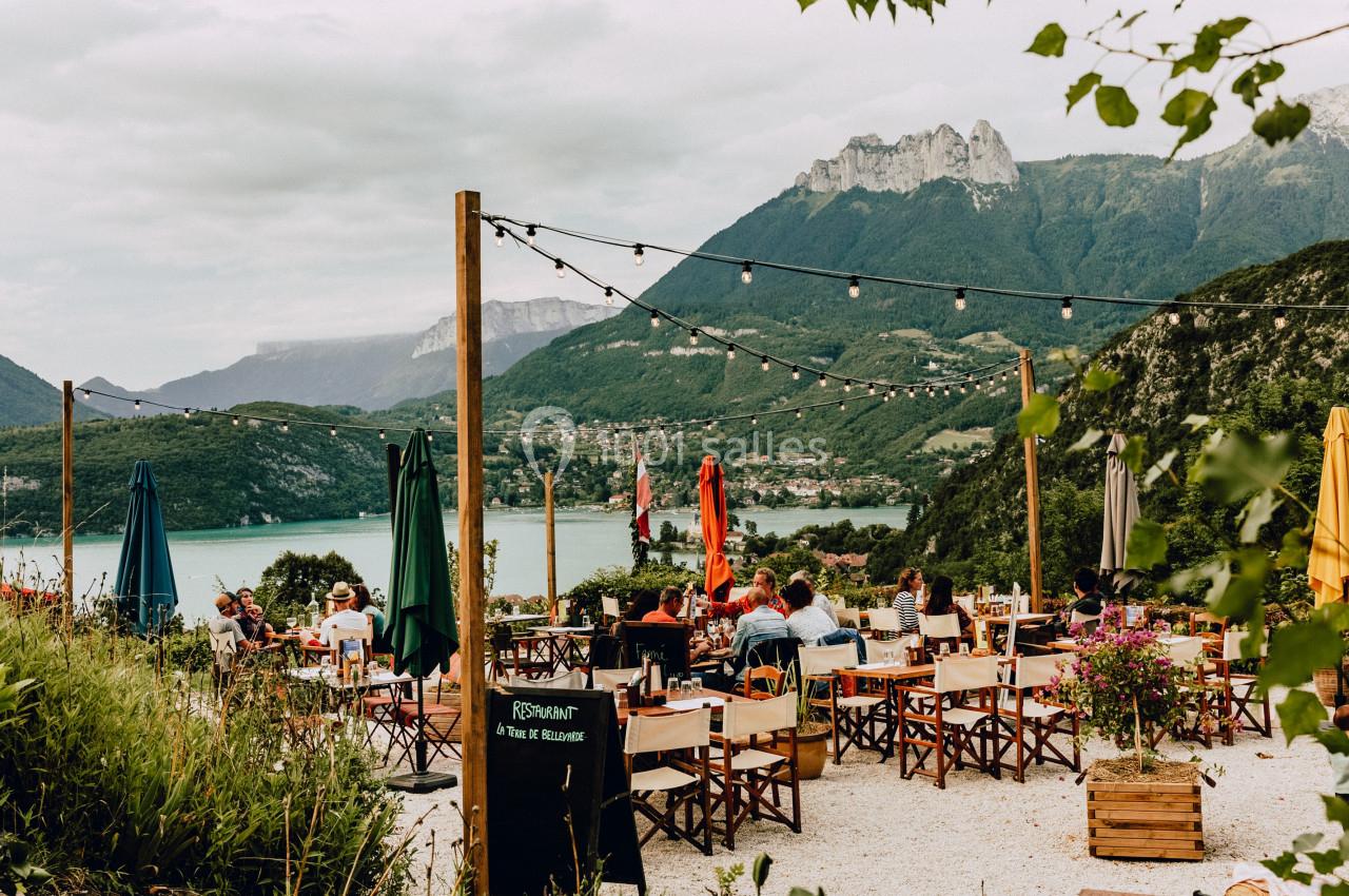 Terrasse d'un restaurant avec vue sur un lac entouré de montagnes, décorée de guirlandes lumineuses.