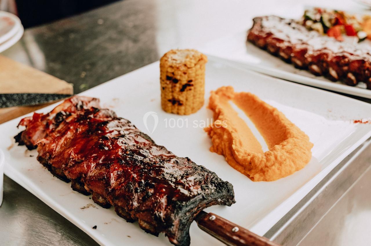 Assiette contenant des côtes de porc grillées, une purée orange, un épi de maïs et des légumes en arrière-plan.
