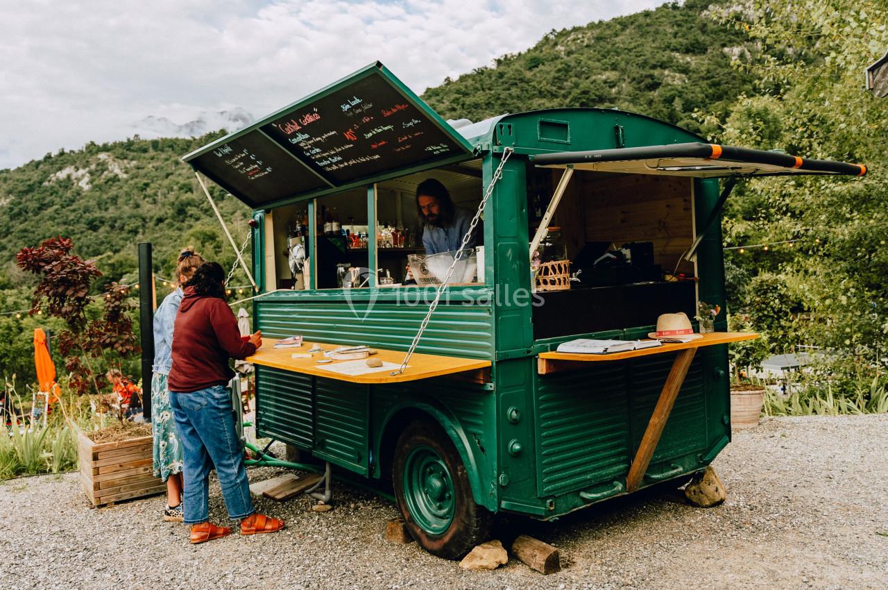 Camion-restaurant vert en plein air avec des clients commandant à un comptoir, entouré de végétation et de collines.