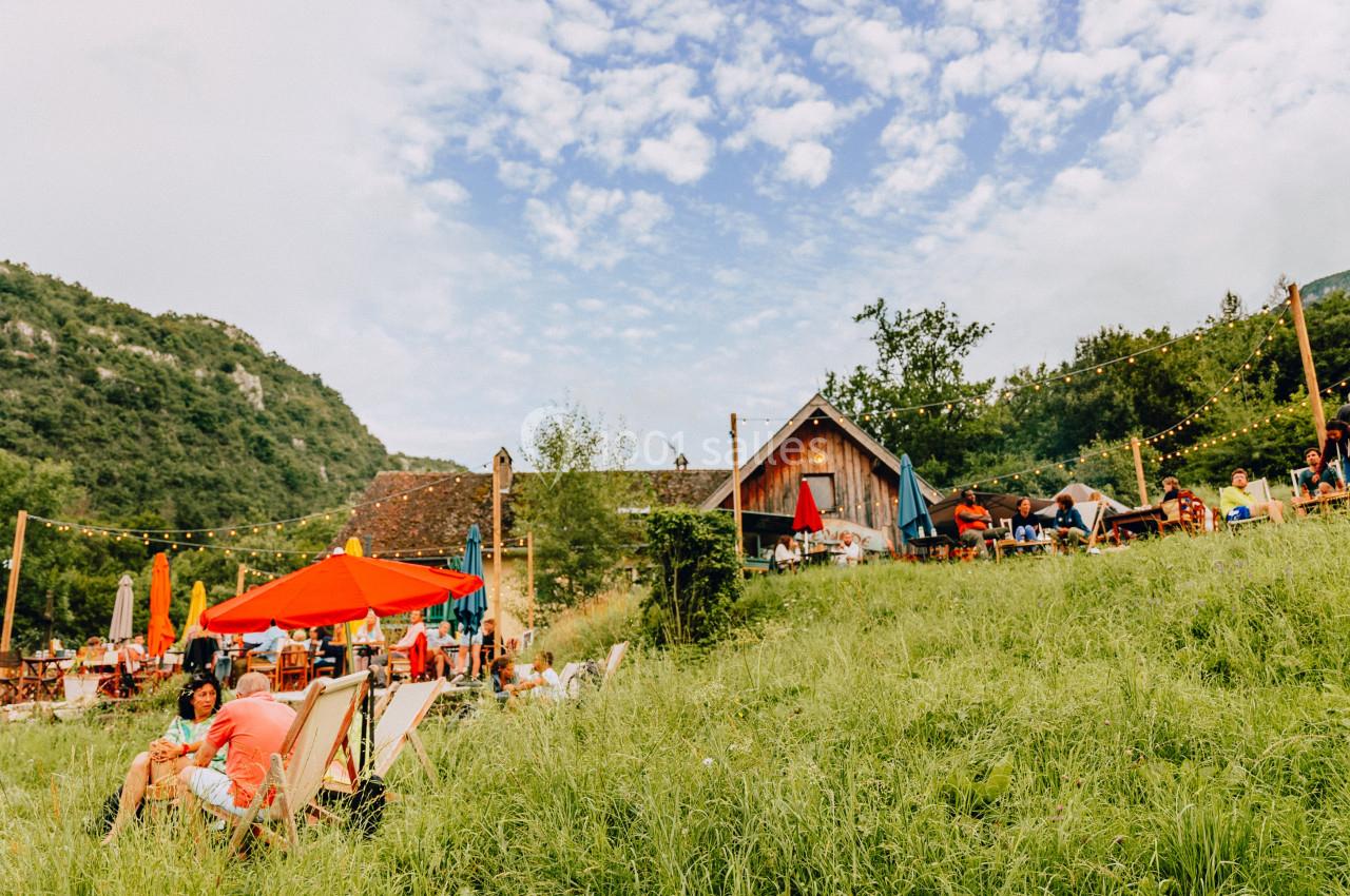Des personnes se détendent sur des transats et à des tables en plein air, entourées de verdure et de collines.