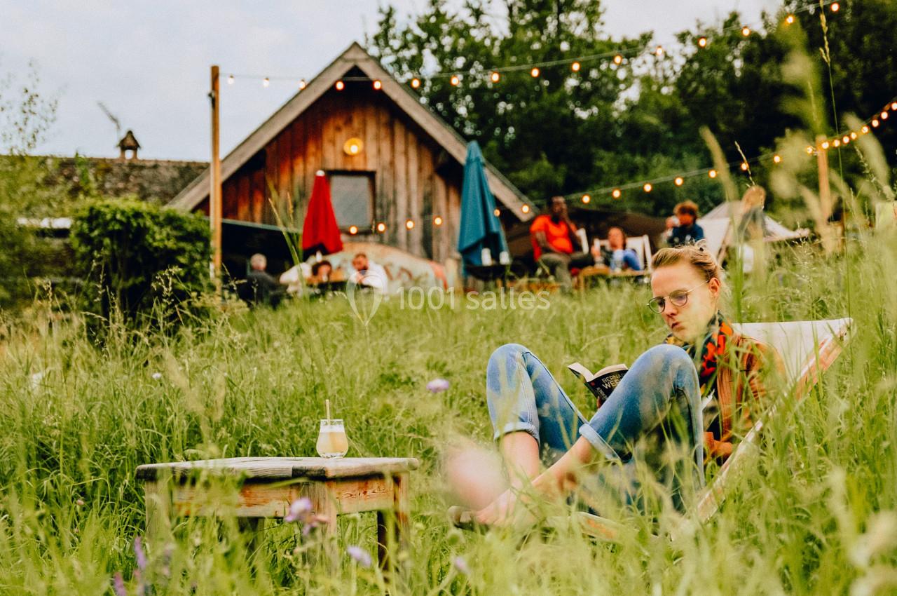 Une femme assise dans un champ verdoyant près d'une table basse, avec une maison en bois et des guirlandes lumineuses en…