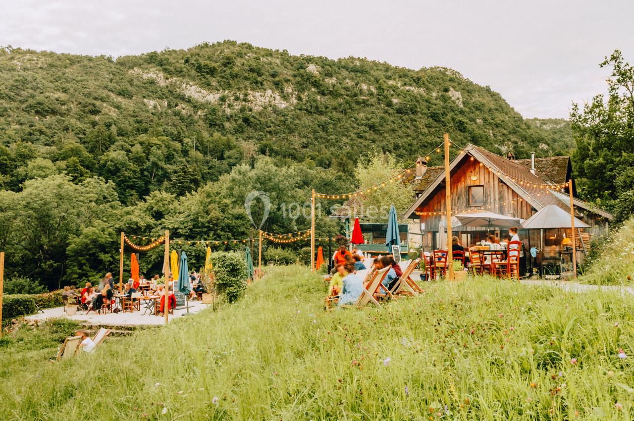 Terrasse d'un café en plein air avec des parasols colorés, située dans un cadre verdoyant au pied de collines boisées.