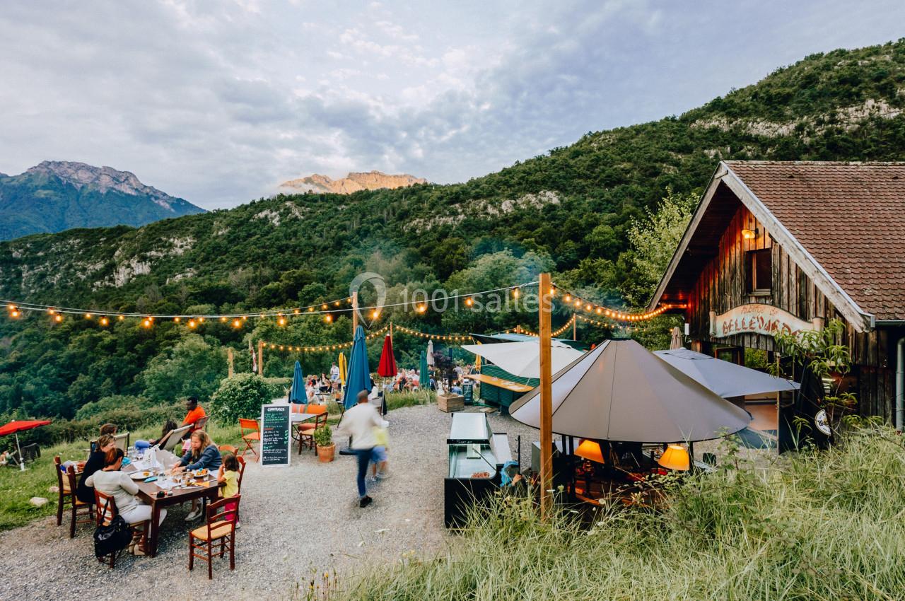 Terrasse extérieure d'un restaurant rural avec des tables, des parasols et des guirlandes lumineuses, entourée de montagnes.
