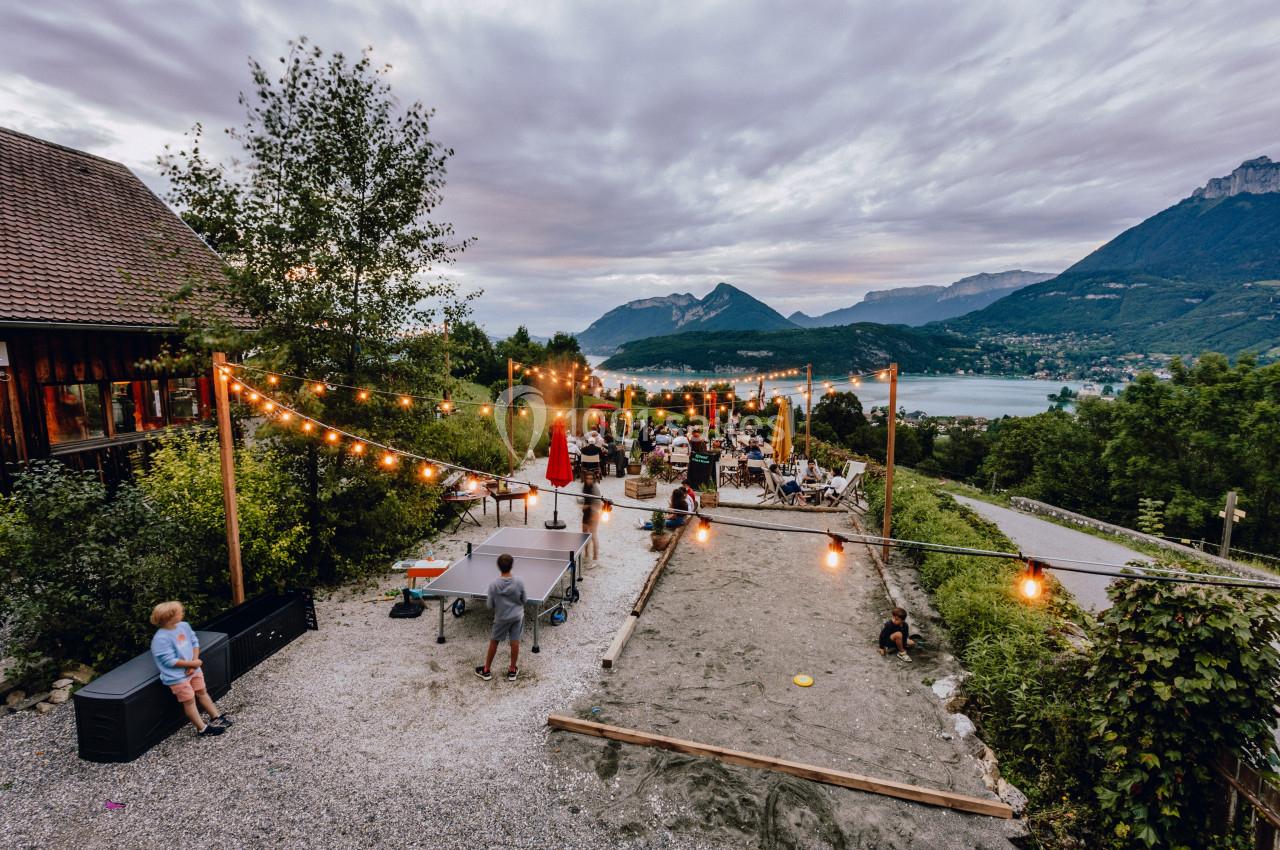 Terrasse extérieure avec guirlandes lumineuses, tables occupées, vue sur un lac et montagnes en arrière-plan.