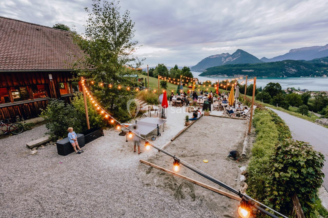 Terrasse extérieure avec guirlandes lumineuses, tables occupées et vue sur un lac entouré de montagnes.