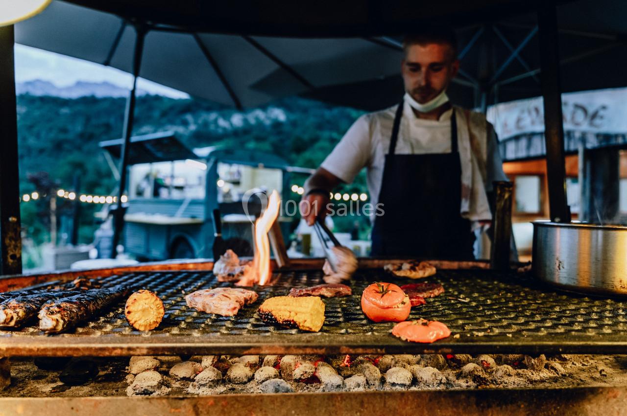 Un cuisinier grille des viandes et des légumes sur un barbecue en plein air, avec des lumières et un paysage en arrière-plan.