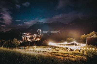 Vue nocturne d'un paysage montagneux avec un village éclairé au loin et une terrasse illuminée au premier plan.