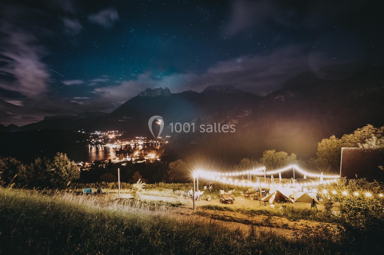 Vue nocturne d'un paysage montagneux avec un village éclairé au loin et une terrasse illuminée au premier plan.