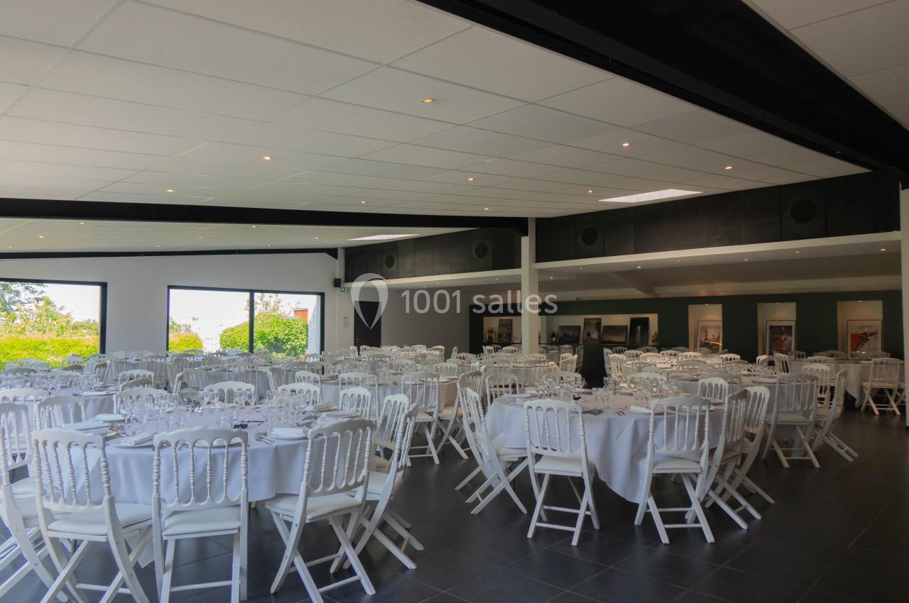 Salle de réception lumineuse avec des tables rondes dressées et des chaises blanches, vue sur l'extérieur verdoyant.