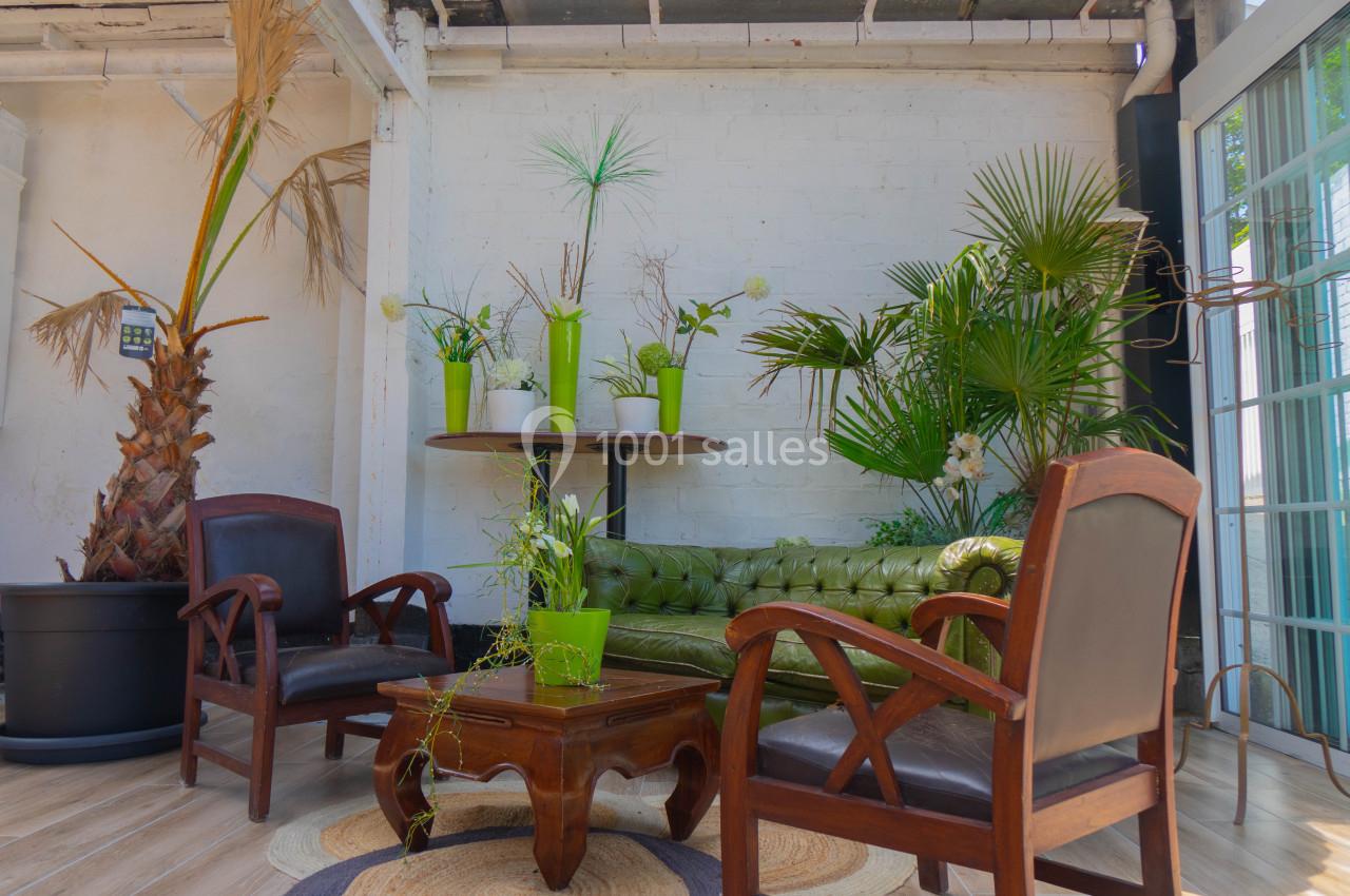 Salon lumineux avec canapé vert, fauteuils en bois, table basse et plantes décoratives contre un mur blanc.