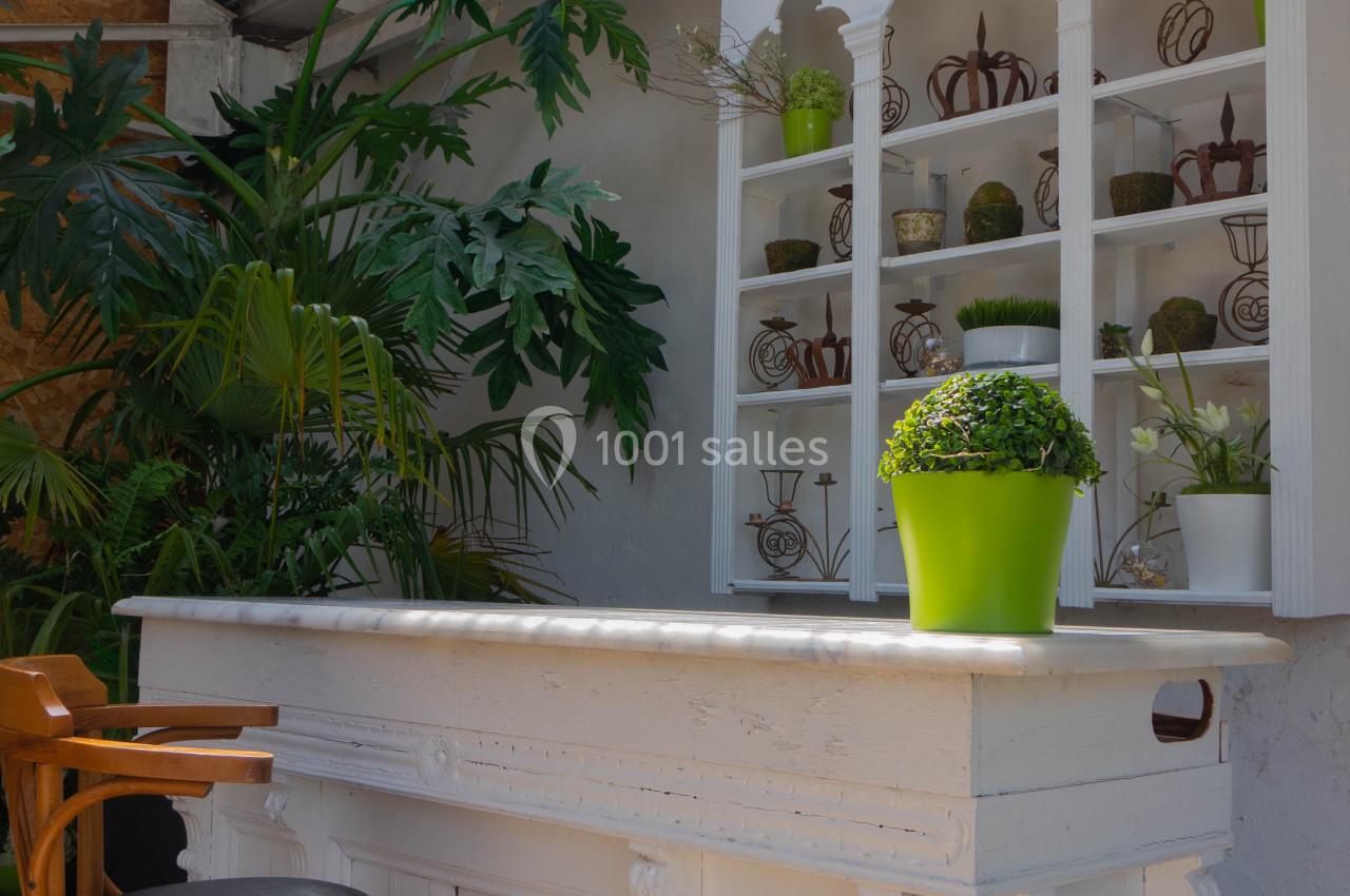 Comptoir blanc avec tabouret en bois, étagère décorative en arrière-plan et plantes vertes sous un toit partiellement ouvert.