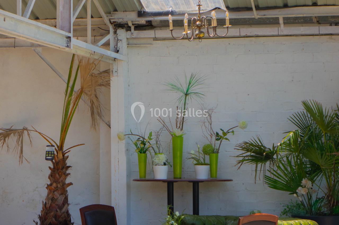 Espace lumineux avec toit en verre, plantes vertes, canapé vert et chaises en bois autour d'une table basse.