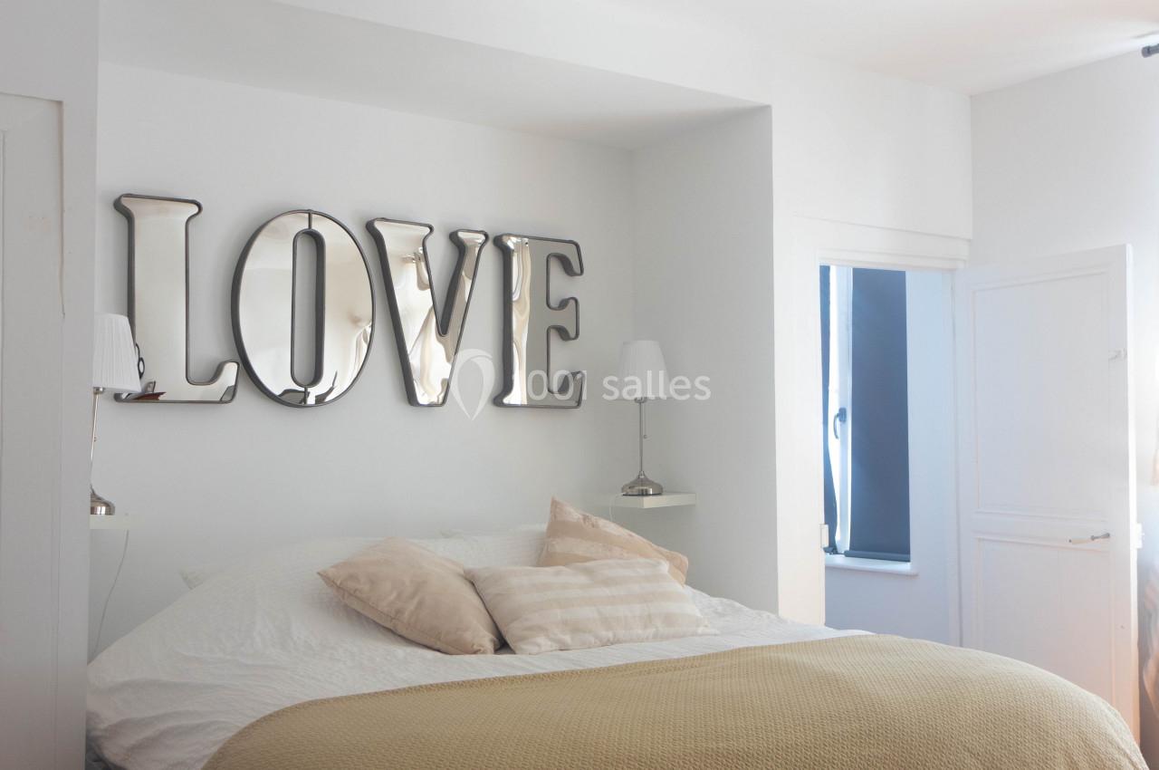 Chambre lumineuse avec un lit double, coussins beiges, parquet en bois et décoration murale ’LOVE’ en lettres métalliques.