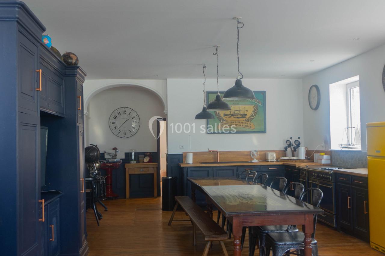 Cuisine spacieuse avec meubles bleu foncé, table en bois, chaises métalliques, réfrigérateur jaune et horloge murale.
