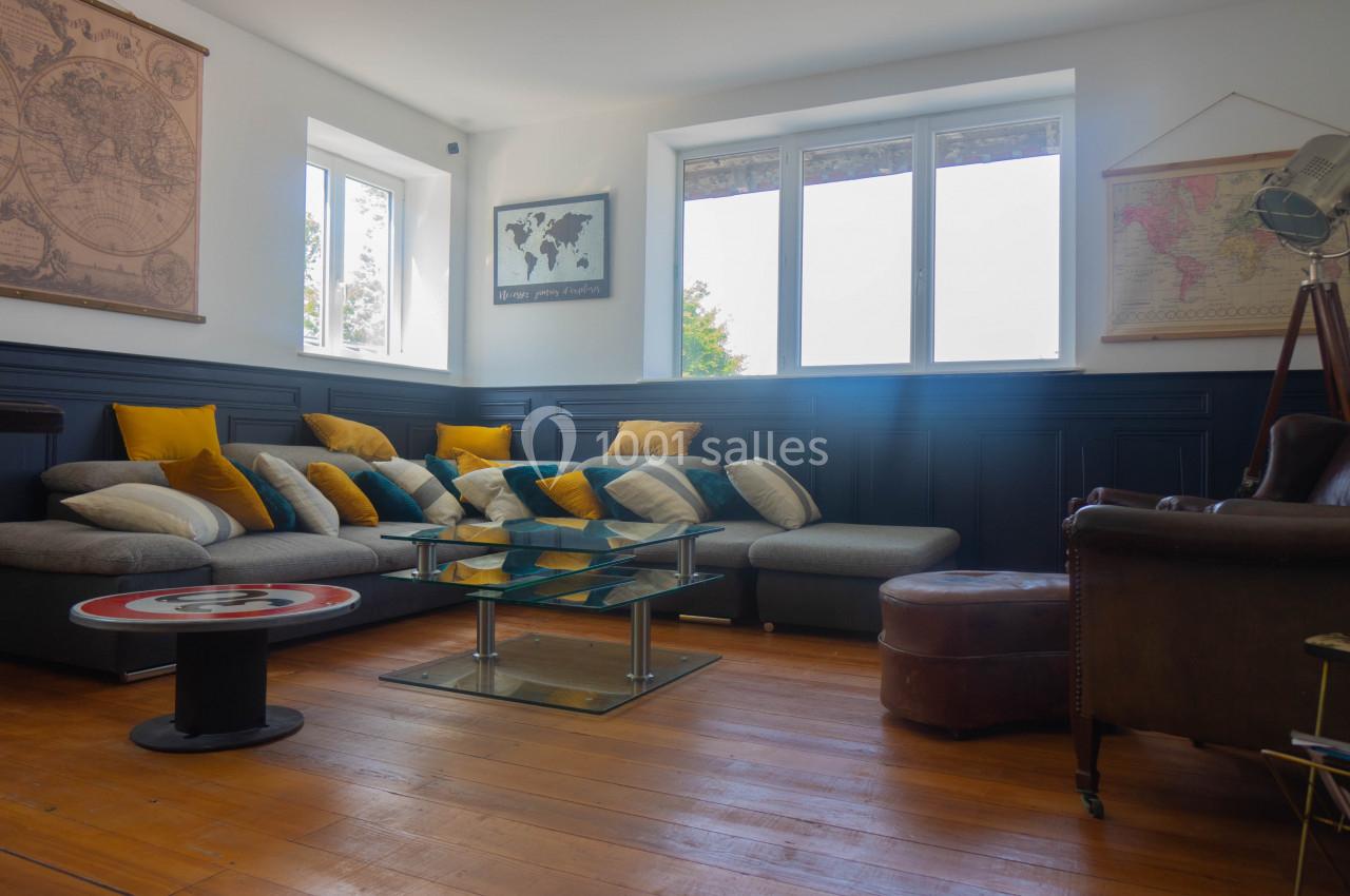 Salon lumineux avec canapé d'angle gris, coussins colorés, table basse en verre et cartes du monde aux murs.