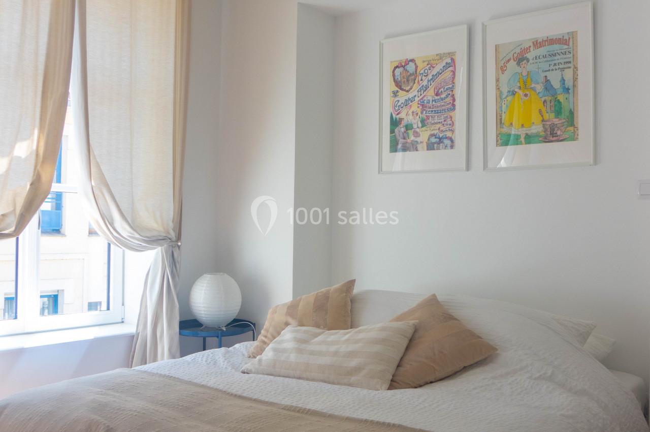 Chambre lumineuse avec lit double, coussins beiges, rideaux clairs et deux cadres accrochés au mur blanc.