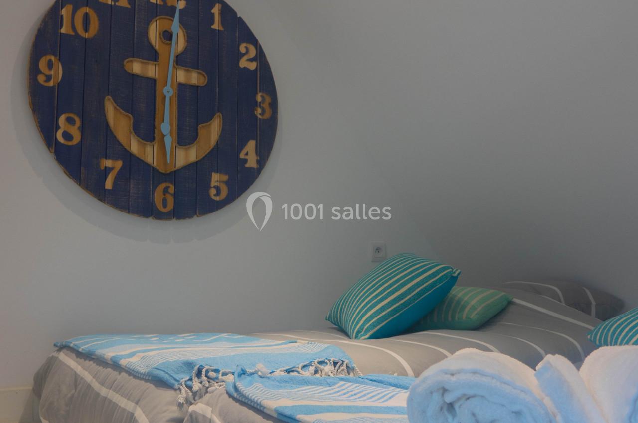 Deux lits simples avec draps bleus et gris, serviettes roulées, et une horloge murale en bois décorée d'une ancre.